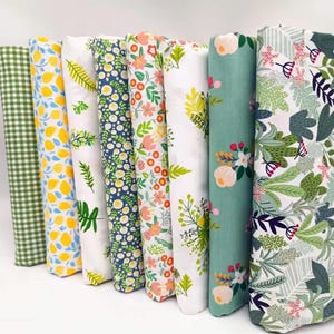 May include: A collection of rolled fabric bolts featuring various floral and patterned designs. The fabrics display a range of colors, including green, yellow, blue, and pink, with patterns such as lemons, daisies, and leaves. The fabrics are suitable for sewing and crafting projects.