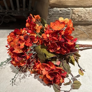 May include: A bouquet of artificial orange and red hydrangeas with green leaves and stems. The flowers are arranged in a natural, full display, perfect for home decor or seasonal arrangements. The bouquet is set against a neutral background.