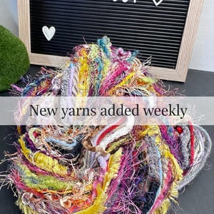 May include: A colorful pile of yarn in various textures and hues, including yellow, pink, and blue. The image features a black letter board with the words "INNER JOY" and a heart symbol. Text overlay reads "New yarns added weekly".