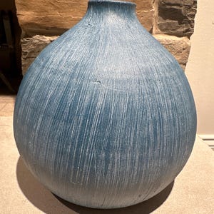 May include: A large, blue decorative vase with a rounded body and a narrow neck. The vase has a textured surface with vertical white lines. The vase is made of ceramic or a similar material and is suitable for home decor.