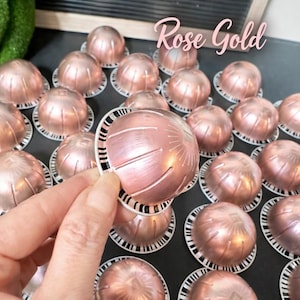 May include: Rose gold coffee capsules with a textured surface and a white and black patterned rim. The image shows a hand holding one capsule, with many more capsules in the background. The text "Rose Gold" is written in a cursive font.