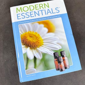 May include: A book titled "Modern Essentials" with a daisy image and three small amber essential oil bottles. The book's subtitle reads "A Contemporary Guide to the Therapeutic Use of Essential Oils". The book is the seventh edition.