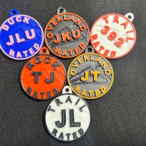 Könnte beinhalten: Sechs runde, schwarz-weiße, plastikartige Schlüsselanhänger mit unterschiedlichen Hintergrundfarben und Texten. Die Anhänger zeigen "DUCK JLU RATED", "OVERLAND JKU RATED", "TRAIL 392 RATED", "ROCK TJ RATED", "OVERLAND JT RATED" und "TRAIL JL RATED".