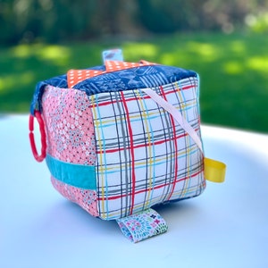 May include: A multi-colored fabric cube with different textures and patterns, including a red and white plaid, a pink floral print, and a blue and white geometric pattern. The cube has a red plastic ring, a turquoise ribbon, and a yellow ribbon attached to it.