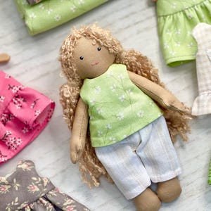 May include: A handmade cloth doll with curly blonde hair, wearing a green floral top and white shorts. Several miniature dresses and skirts in various floral patterns and colors surround the doll, suggesting a clothing set.