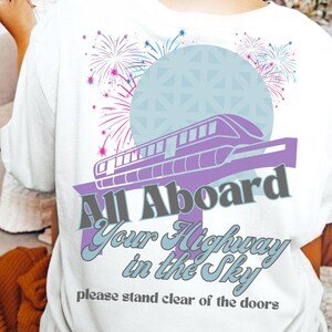 May include: White t-shirt with a purple and gray graphic of a monorail with the text "All Aboard Your Highway in the Sky" and "please stand clear of the doors".