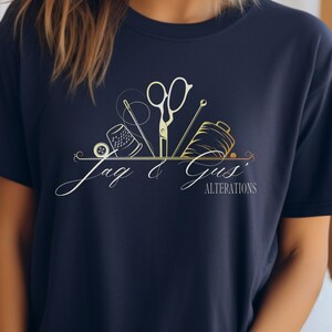 May include: A navy blue t-shirt with a gold and white logo that says "Jag & Gus' Alterations". The logo features a sewing needle, thread, thimble, and scissors.