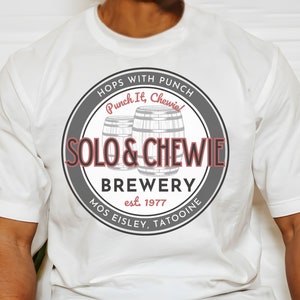 Puede incluir: Camiseta blanca con un logotipo circular con fondo gris, dos barriles de cerveza grises y el texto "HOPS WITH PUNCH", "Punch It, Chewie", "SOLO & CHEWIE", "BREWERY", "est. 1977", "MOS EISLEY, TATOOINE" en blanco.