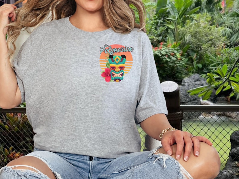 Puede incluir: Una camiseta gris con un gr&aacute;fico de una m&aacute;scara tiki con una flor roja y el texto "The Polynesian" en una fuente retro.