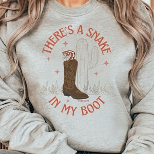 May include: A light grey sweatshirt with a graphic design. The design features a brown cowboy boot with a snake, a cactus, and the text "THERE'S A SNAKE IN MY BOOT" in a rust colour. The text "ESTD 1995" is also included.