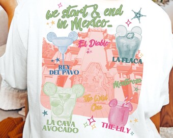 May include: A white t-shirt with a colorful graphic design featuring a pink and orange castle with Mickey Mouse ears. The text "we start & end in Mexico" is written in green. The graphic also includes four cocktails with names like "La Flaca" and "The Lily".