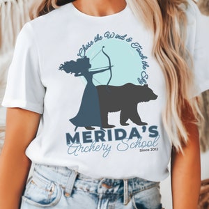 May include: White t-shirt with a graphic of a woman with a bow and arrow, a bear, and the text "Chase the Wind & Touch the Sky." Below, it reads "Merida's Archery School Since 2012."