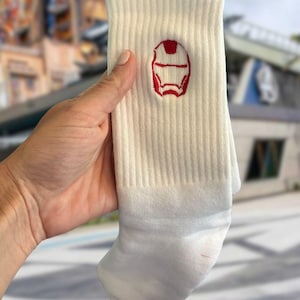 May include: Cream-colored socks with a red embroidered Iron Man mask design. The socks are ribbed and have a white toe and heel. The design is a stylized outline of the superhero's helmet.