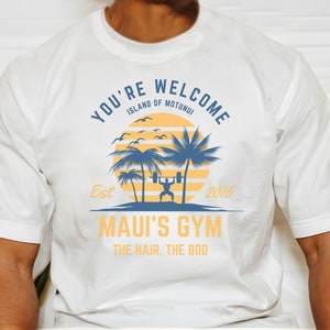 May include: White t-shirt with a blue and yellow graphic design. The graphic features a sunset, palm trees, a person lifting weights, and the text "You're Welcome Island of Motunui Est 2016 Maui's Gym The Hair, The Bod".