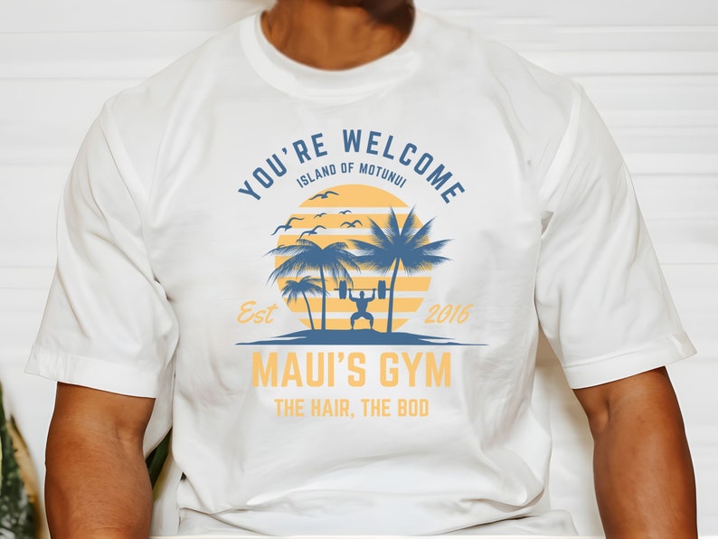 May include: White t-shirt with a blue and yellow graphic design. The graphic features a sunset, palm trees, a person lifting weights, and the text "You're Welcome Island of Motunui Est 2016 Maui's Gym The Hair, The Bod".