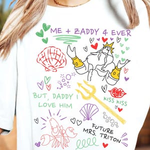 Puede incluir: Camiseta blanca con dibujos de dibujos animados coloridos. El dise&ntilde;o incluye una concha, corazones, un tridente y el texto "ME + ZADDY 4 EVER", "BUT, DADDY I LOVE HIM", "KISS KISS" y "FUTURE MRS. TRITON".