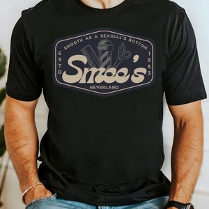 May include: Black t-shirt with a vintage-style logo for a barbershop called "Smee's" in a tan color. The logo includes a barber pole, scissors, and a razor. The text "Smooth as a Seagull's Bottom" is above the logo, and "Est. 1953 Neverland" is below the logo.