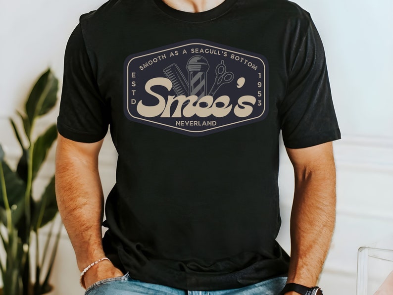 May include: Black t-shirt with a vintage-style logo for a barbershop called "Smee's" in a tan color. The logo includes a barber pole, scissors, and a razor. The text "Smooth as a Seagull's Bottom" is above the logo, and "Est. 1953 Neverland" is below the logo.