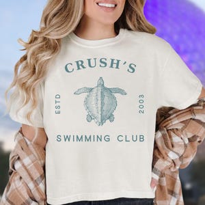 May include: White t-shirt with a blue graphic of a sea turtle and the text "Crush's Est'd 2003 Swimming Club".