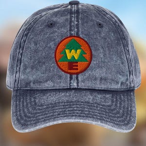 Puede incluir: Gorra de béisbol de color denim con visera curva. La gorra presenta un parche bordado con un árbol verde, una "W" amarilla y detalles rojos y marrones. Botón azul oscuro en la parte superior.