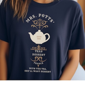 Puede incluir: Una camiseta azul marino con un gráfico de tetera blanca y el texto "MRS. POTTS, ESTD 1991 TEA& DESSERT FRANCE WITH THE TEA, SHE'LL WANT DESSERT".