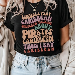 May include: Black t-shirt with a colorful graphic that says "I usually say Caribbean unless I'm talking about Pirates of the Caribbean then I say Caribbean".