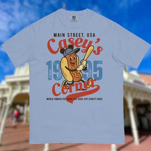 May include: Light blue t-shirt featuring a cartoon hot dog character wearing a baseball cap and holding a bat. The shirt has the text "Main Street, USA Casey's Corner 1995" in red and blue. The bottom of the shirt reads "World Famous Hot Dogs, Hot Diggity Dogs."
