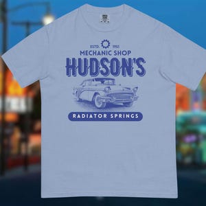 May include: Pale blue t-shirt with a classic car illustration. The shirt features the text "HUDSON'S MECHANIC SHOP" and "RADIATOR SPRINGS" below. The design includes the year of establishment, 1951.