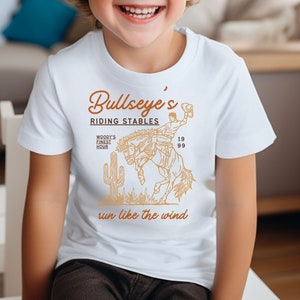 May include: White t-shirt featuring a brown graphic of a cowboy riding a horse. The graphic includes the text "Bullseye's Riding Stables", "Woody's Finest Hour", "19 99", and "run like the wind".