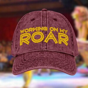 May include: A maroon baseball cap with the text "WORKING ON MY ROAR" embroidered in yellow. The cap has a curved brim and a distressed, vintage look. The text is in a bold, outlined font.