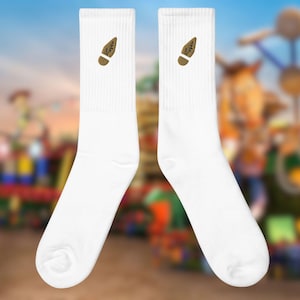 Puede incluir: Dos calcetines blancos con un diseño de huella marrón en la parte superior. Los calcetines se muestran sobre un fondo borroso de un parque de atracciones colorido, lo que sugiere un tema lúdico. Los calcetines están hechos de un material suave y acanalado.
