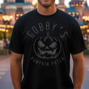 Puede incluir: Una camiseta negra con un gráfico blanco de una calabaza con una cara de miedo y el texto "Gobbly's Pumpkin Patch".