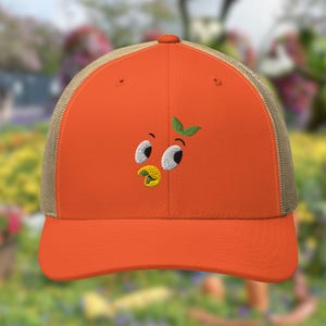 May include: Orange and brown trucker hat with an embroidered orange character with a green leaf on top. The character has large eyes and a yellow beak.