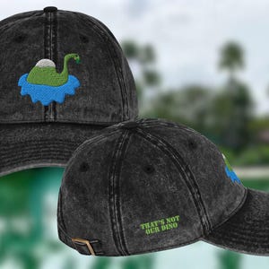 May include: Black baseball cap with a distressed finish. The cap features an embroidered design of a green dinosaur in a blue lake. The text "THAT'S NOT OUR DINO" is embroidered on the back.