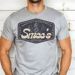 May include: A gray t-shirt with a navy blue logo that says "Smooth as a Seagull's Bottom" and "Smee's" with the text "Est'd 1953" and "Neverland" underneath. The logo also features a barber pole, a razor, and a pair of scissors.