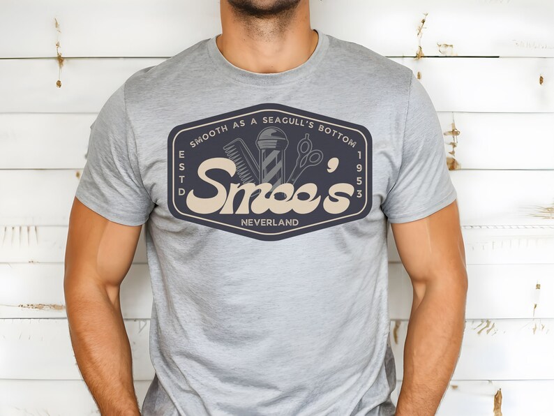 May include: A gray t-shirt with a navy blue logo that says "Smooth as a Seagull's Bottom" and "Smee's" with the text "Est'd 1953" and "Neverland" underneath. The logo also features a barber pole, a razor, and a pair of scissors.
