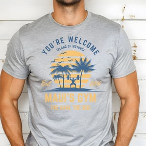 May include: Gray t-shirt with a graphic design of a palm tree and a person lifting weights. The text on the shirt reads "You're Welcome Island of Motunui Est 2016 Maui's Gym The Haul The Dog".