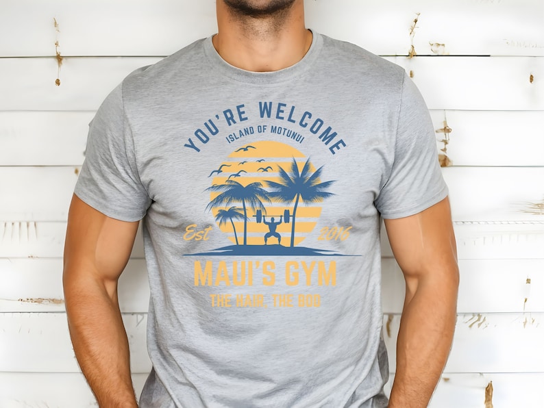 May include: Gray t-shirt with a graphic design of a palm tree and a person lifting weights. The text on the shirt reads "You're Welcome Island of Motunui Est 2016 Maui's Gym The Haul The Dog".
