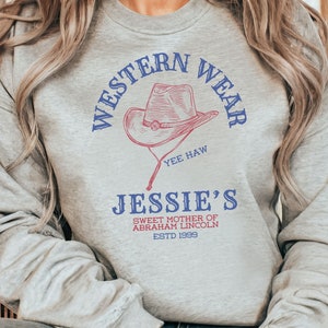 May include: Grey sweatshirt with the text "Western Wear" in blue and red lettering, a drawing of a cowboy hat, and the text "Jessie's Sweet Mother of Abraham Lincoln Est'd 1999" in blue lettering.