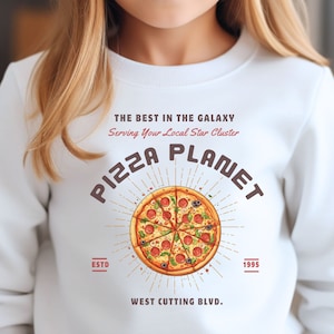 K&ouml;nnte beinhalten: Wei&szlig;er Pullover mit einem Grafikdesign einer Pizza mit dem Text "Pizza Planet" in einer Retro-Schrift. Der Text "The Best in the Galaxy" und "Serving Your Local Star Cluster" befindet sich &uuml;ber dem Pizzagrafi. Der Text "ESTD" und "1995" befindet sich auf beiden Seiten des Pizzagrafiks. Der Text "West Cutting Blvd." befindet sich unter dem Pizzagrafi.