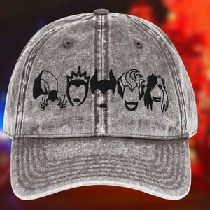 May include: Grey baseball cap with a distressed finish. The cap features a black graphic of four stylized villain faces across the front. The brim is curved, and the cap has a button on top.