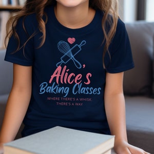 May include: Navy blue t-shirt with the text "Alice's Baking Classes" in pink and blue script. The design includes a whisk, rolling pin, and a heart. The shirt also has the phrase "Where there's a whisk, there's a way."
