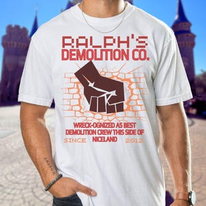May include: White t-shirt with the text "RALPH'S DEMOLITION CO." in red, featuring a brown fist breaking through a brick wall graphic. The text "WRECK-OGNIZED AS BEST DEMOLITION CREW THIS SIDE OF NICELAND SINCE 2012" is also printed on the shirt.