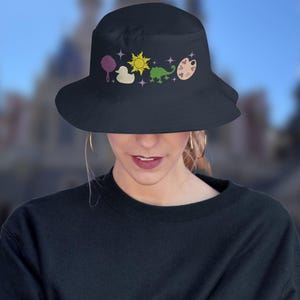 May include: A black bucket hat with a design of various Disney princess icons, including a frying pan, a duck, a sun, a chameleon, and a paint palette.