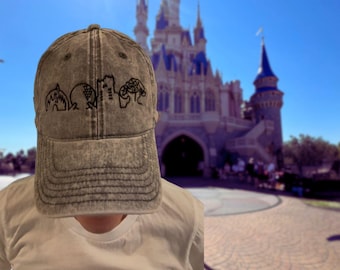 Könnte beinhalten: Eine graue Baseballkappe aus Denim mit schwarzen gestickten Symbolen von vier Disney-Themenpark-Attraktionen. Die Kappe wird von einer Person vor einem verschwommenen Hintergrund von Cinderellas Schloss in Walt Disney World getragen.