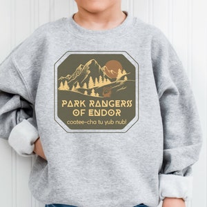 May include: Gray sweatshirt with a brown and yellow graphic design featuring a mountain range, trees, and a sun. The text reads "Park Rangers of Endor" and "coatee-cha tu yub nub!"
