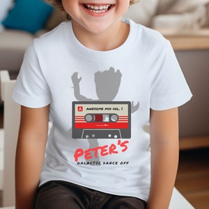 Puede incluir: Camiseta blanca con una silueta gris de una figura bailando sosteniendo una cinta de casete con el texto "Awesome Mix Vol. 1" y "Peter's Galactic Dance Off".