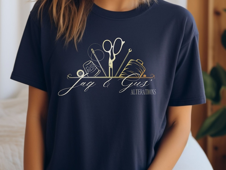 May include: A navy blue t-shirt with a gold and white logo that says "Jag & Gus' Alterations". The logo features a sewing needle, thread, thimble, and scissors.