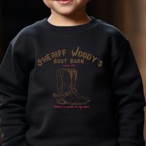 Puede incluir: Sudadera negra con el texto "SHERIFF WOODY'S BOOT BARN" y una ilustraci&oacute;n de bota de vaquero. Tambi&eacute;n incluye el texto "Since 1995" y "There's a snake in my boot". El dise&ntilde;o es marr&oacute;n y dorado.