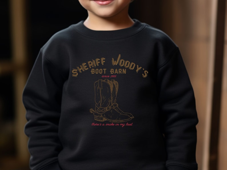 Puede incluir: Sudadera negra con el texto "SHERIFF WOODY'S BOOT BARN" y una ilustraci&oacute;n de bota de vaquero. Tambi&eacute;n incluye el texto "Since 1995" y "There's a snake in my boot". El dise&ntilde;o es marr&oacute;n y dorado.